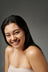 positive and young asian woman with brunette hair and acne prone skin smiling on grey background