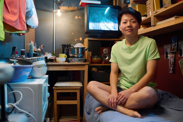 Young Asian woman in casualwear sitting on bed in small apartment and looking at camera against old...