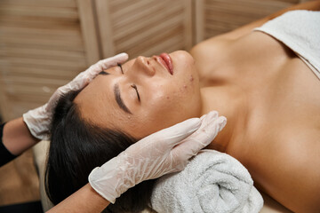 facial massage for acne-prone skin, therapist in latex gloves and asian client with closed eyes