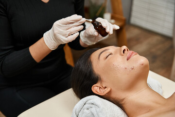 Obraz na płótnie Canvas therapist applying treatment serum with cotton swab on face of asian woman with acne-prone skin