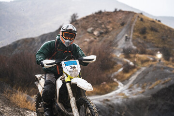 Professional motorcyclist in full moto equipment riding crops enduro bike, portrait of an enduro rider at a competition in the mountains, rainy and cloudy weather, concept of motosport