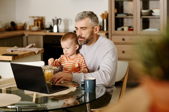 Serious mature male solopreneur with his baby son on knees sitting on chair by kitchen table and looking at laptop screen during network