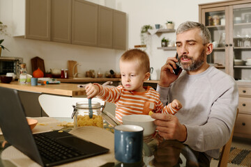 Confident mature businessman with baby son on his knees talking to colleague or subordinate on...