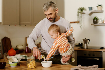 Mature man with cute baby son on hands pouring fresh milk into bowl with cornflakes while preparing...