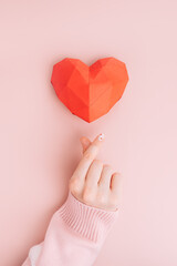 Hand shows heart sign under red origami heart shape on pink background.