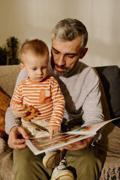 Mature father with open book of comics and adorable baby son pointing at page with picture sitting on couch in front of camera