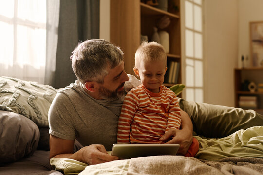 Mature father with tablet looking at smiling baby son while both relaxing on bed or couch in front of camera and watching online cartoon