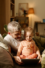 Mature father and his adorable baby son relaxing on bed or couch in front of camera and watching online cartoon on screen of tablet