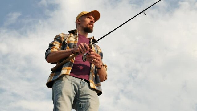 Looking up, a young fisherman in a shirt and a cap catches fish using a spinning rod with a fishing reel, throwing bait into the water, swinging a fishing rod, active lifestyle. Hobbies and interests