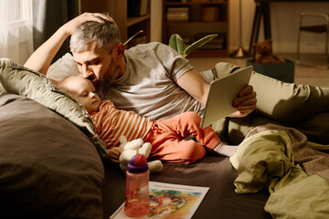 Man with grey hair kissing his adorable baby son lying next to him and looking at screen of tablet...