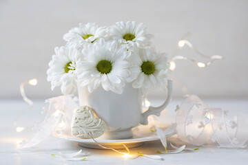 White daisy chrysanthemum flowers in a cup, light garland, heart and ribbon on a white wooden table. Beautiful card for the holiday.