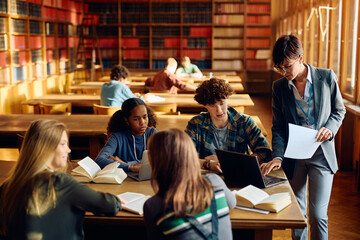 High school teacher using laptop with her students in library.