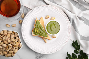 Tasty pistachio cream in bowl, nuts and toasts on white marble table, flat lay
