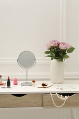 Mirror, cosmetic products, jewelry and vase with pink roses on white dressing table in makeup room