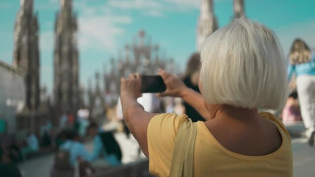 Happy senior woman tourist taking selfie photos or video for her travel blog outdoors in front of a roof view of famous statue on cathedral Duomo di Milano Cathedral