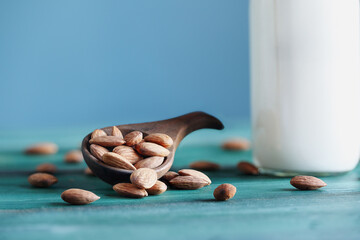 Close up of whole fresh almonds in a wooden spoon with a bottle of fresh creamy almond white milk over a rustic table or background. Selective focus with blurred background.