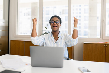Overjoyed excited african-american woman celebrating victory sitting in the coworking space in...