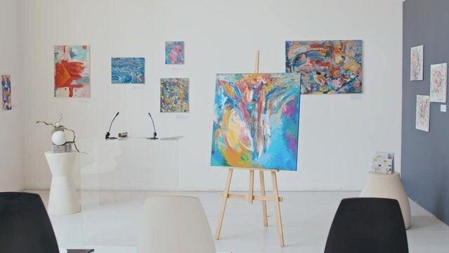Full pan no people shot of interior of modern art gallery during exhibition, with expressionist and abstract oil and acrylic paintings on easel and white walls, in the morning before opening