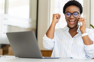 Happy excited african-american woman celebrating victory sitting in office in front of the laptop, talking on the smartphone, lucky businessman screaming yes, receved good news by phone, job offer