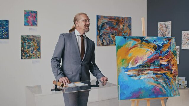 Medium full shot of mature Caucasian male art museum director talking in front of press and flashing cameras, and presenting new contemporary abstract painting purchased for collection
