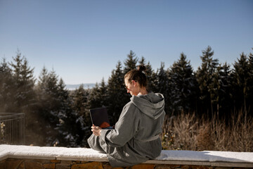 Fototapeta premium Frau im Bademantel sitzt auf einer verschneiten Steinmauer in der Natur und arbeitet am laptop