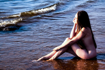Pretty naked naturist woman sitting on nudist public sea beach in water, looking away, relaxing. Lovely nude lady with sexy body. Nudism naturism lifestyle concept, clothing optional. Copy text space - obrazy, fototapety, plakaty
