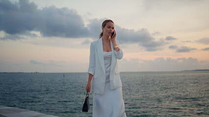 Elegant lady calling at sunset sky. Woman talking smartphone walking embankment