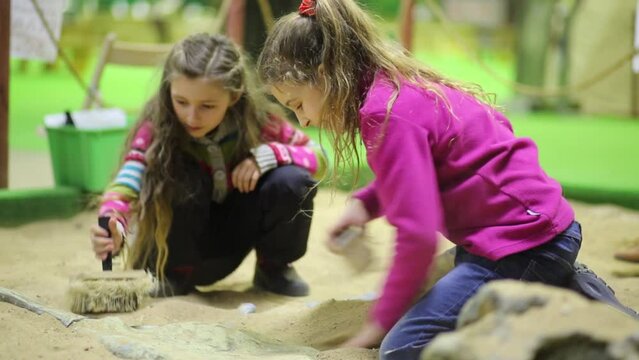Two girls with brushes dig into sand dinosaur bones together