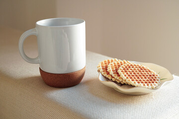 Breakfast of a coffee in a white mug with a cork bottom and Italian waffles on a white sofa lit by the morning light.