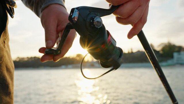 Man hobby fishing on sea tightens fishing line reel of fish summer. Handle rotation with reel of fishing rod against of orange sunset slow motion. Lens flare. Calm surface sea. Bright disk of sun