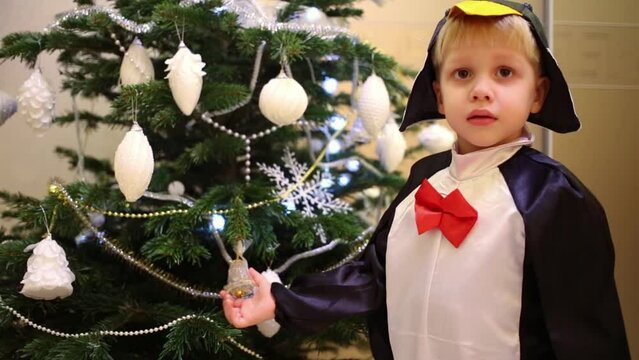 Little boy in penguin costume is holding bells near christmas tree.