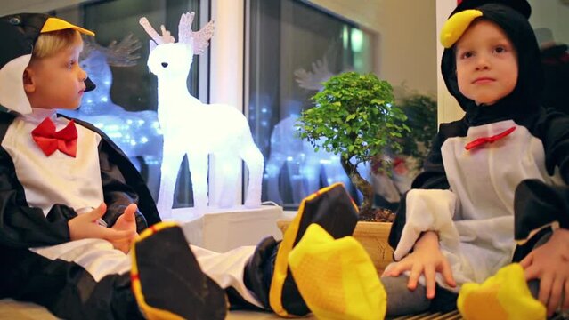 Two little boys in a penguin costume are sitting near deer and tree