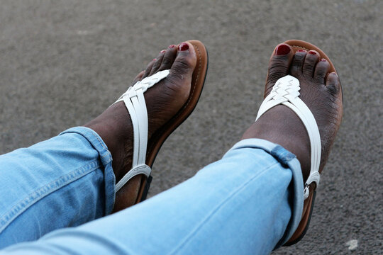 Close-up on the feet of a black woman. Blue jeans. White sandals. Fat feet. Painted nails.