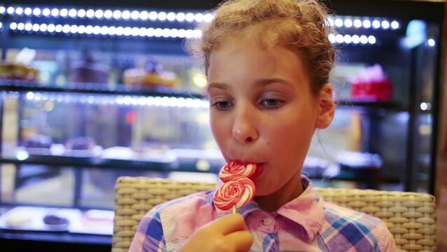 Little girl in shirt licking sweet lollipop at shop