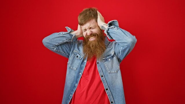 Stressed young redhead man tiredly covering ears over noisy anxiety, isolated on red background, expressing emotional headache problem