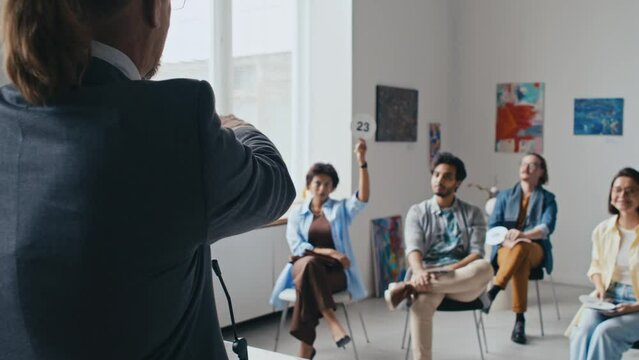 Rear shot of male auctioneer in suit, with ponytail conducting auction in modern art gallery, and multiracial customers sitting on chairs and raising number cards to place bet on paintings on sale