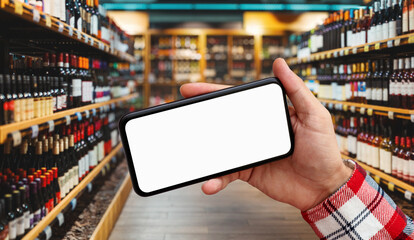 Blank screen of smart phone in male hand in front of wine bottles in liquor store.