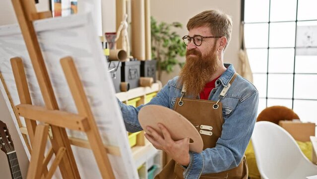 Confident young redhead man rocking the art studio, smiling as he lets his creativity run wild, brush in hand, drawing masterpieces