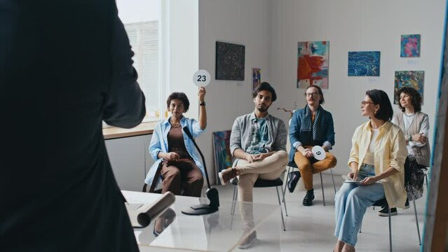 Full rear shot of unrecognizable male salesman conducting auction in contemporary art gallery, and multiethnic buyers sitting on chairs and raising number cards to bet on artworks on sale