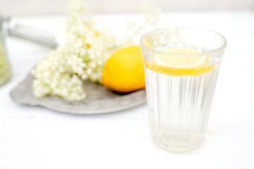 A faceted glass with a slice of lemon near a vase with fresh elderberry flowers on a white background.