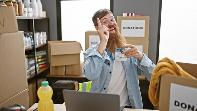 Smiling redhead man volunteering at charity center, earnestly pointing to his identification card while working on his laptop