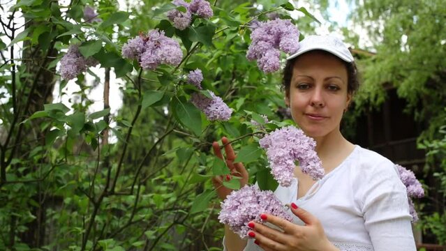 Young woman is standing and smelling the branch of purple lilac.