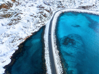 Aerial view of snowy road to the island and blue sea on both side. Bridge on snow and azure transparent water in winter. Landscape. Top drone view of road to the Henningsvaer, Lofoten islands, Norway
