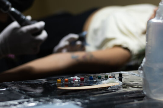 Horizontal photograph of a tattoo artist's workspace where he has his work tools, such as inks, bottles and Vaseline, where in the background you can see a female client getting a tattoo