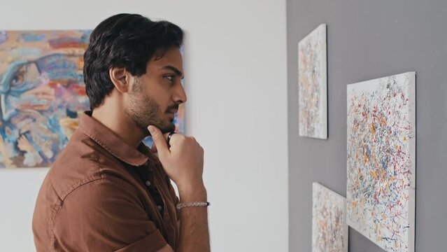 Medium side shot of pensive young Middle Eastern male visitor standing in modern art gallery, looking at abstract artworks on display, leaning in closer to look at detail, stroking beard and nodding