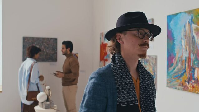 Chest-up arc shot of eccentric Caucasian male visitor with red beard and moustache, in glasses and fedora hat admiring painting at contemporary art exhibition in gallery, couple discussing artworks