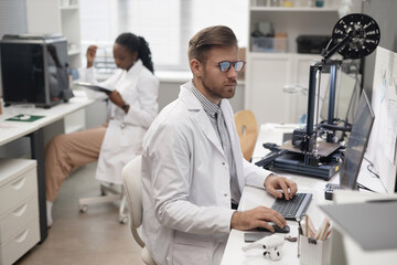 Caucasian man technician in eyeglasses typing on keyboard and looking at computer screen while sitting at desk in 3D printing laboratory, female coworker sitting in defocused background