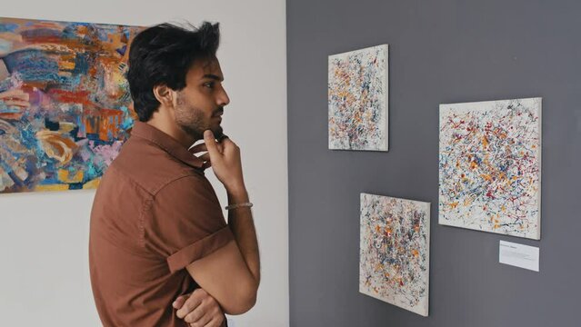 Medium arc shot of thoughtful young Middle Eastern man in casual shirt standing in modern art museum, looking at abstract paintings on display, stroking beard and reading information on label