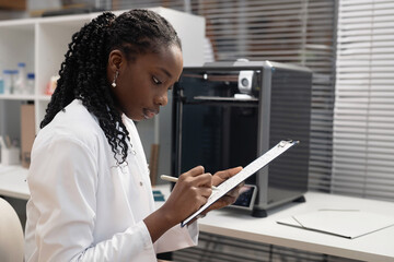 Side view of young African American woman technician filling in document on clipboard in hand while...