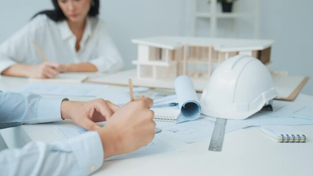 Professional architect engineer drawing blueprint and working together with young beautiful caucasian coworker on meeting table with safety helmet, house model, blueprint scatter around. Immaculate.